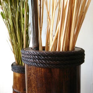 May include: Two brown bamboo vases with a dark brown stain. The vases are decorated with a thick black rope around the top. The vases are filled with dried flowers and branches.