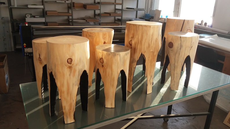 May include: Six wooden stools with natural wood grain and black painted legs. The stools are arranged in a row on a glass table.