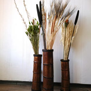 May include: Three tall brown bamboo vases with different dried flowers and grasses. The vases are all the same style and have a dark brown stain.