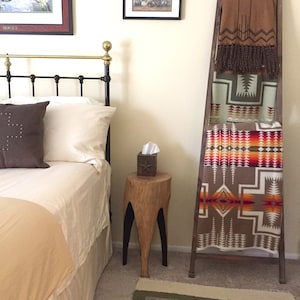 May include: A bedroom with a bed, a wooden ladder with blankets folded on it, a wooden stool with a tissue box on it, and a green rug on the floor.