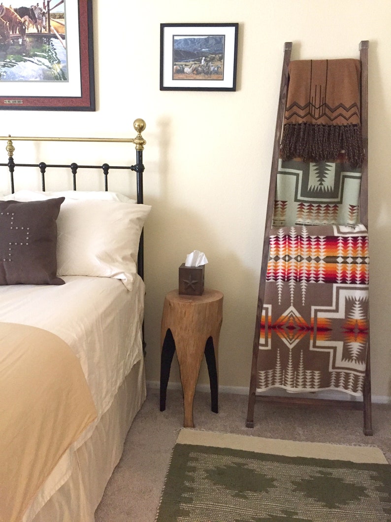 May include: A bedroom with a bed, a wooden ladder with blankets folded on it, a wooden stool with a tissue box on it, and a green rug on the floor.