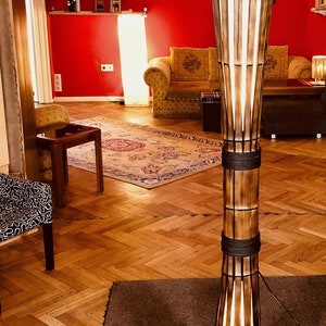Op de afbeelding: Een hoge, houten vloerlamp met een witte kap en een geweven bamboe ontwerp. De lamp staat op een bruin tapijt in een kamer met houten vloer.