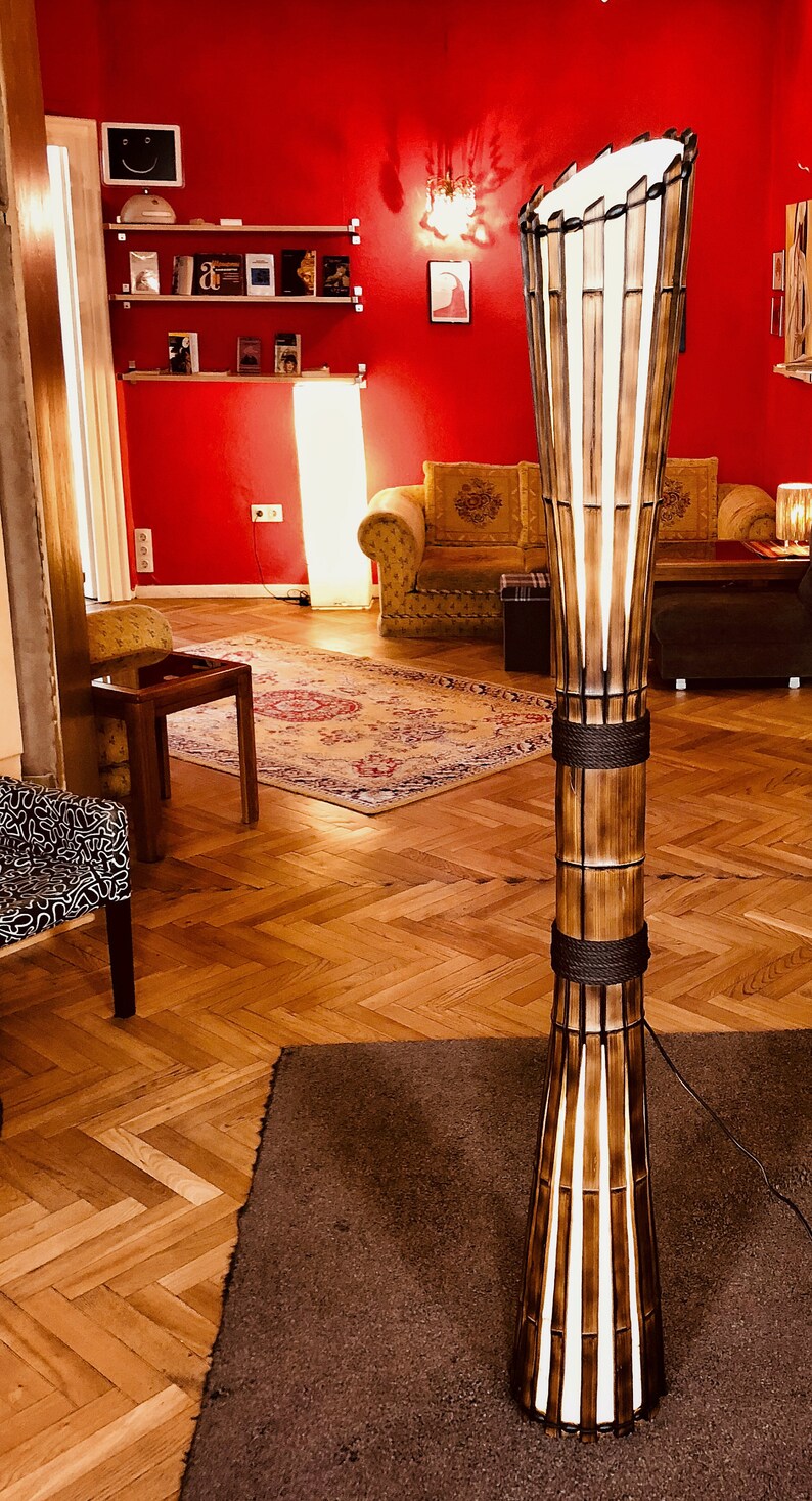 Op de afbeelding: Een hoge, houten vloerlamp met een witte kap en een geweven bamboe ontwerp. De lamp staat op een bruin tapijt in een kamer met houten vloer.