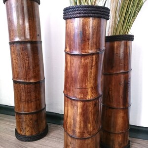 May include: Three tall brown bamboo vases with black rope trim. The vases are empty and standing on a wooden floor.