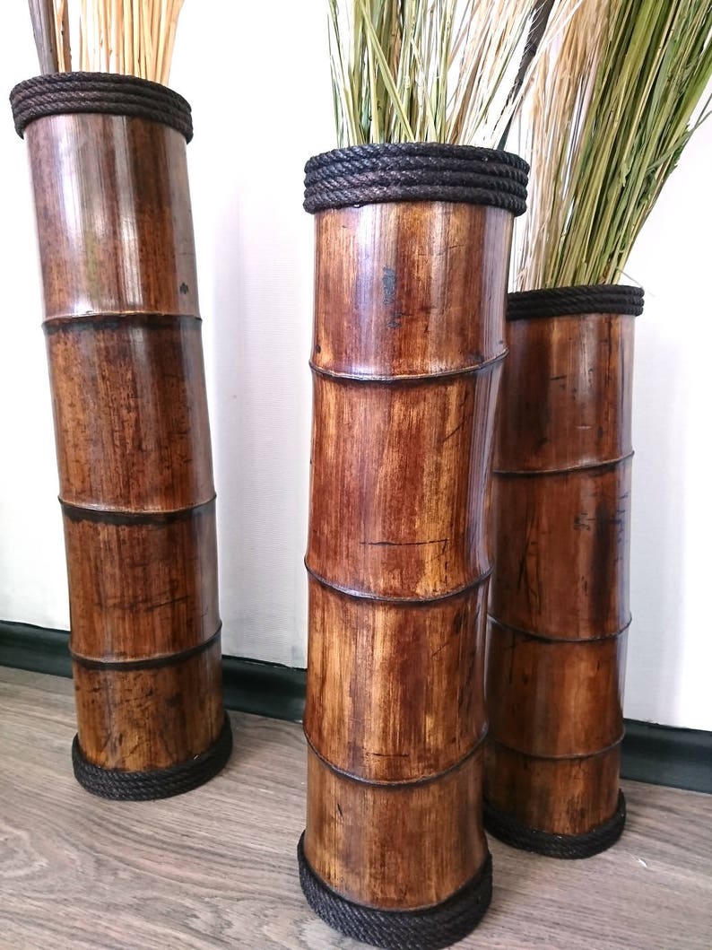 May include: Three tall brown bamboo vases with black rope trim. The vases are empty and standing on a wooden floor.