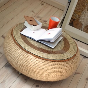 May include: A round, rope-wrapped coffee table with a glass top. The table has a natural brown and beige color scheme. A book and a red cup are on top of the table.