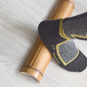 May include: A person's foot wearing a black and gray sock with yellow accents is rolling a round, natural bamboo roller on a light gray wood floor.