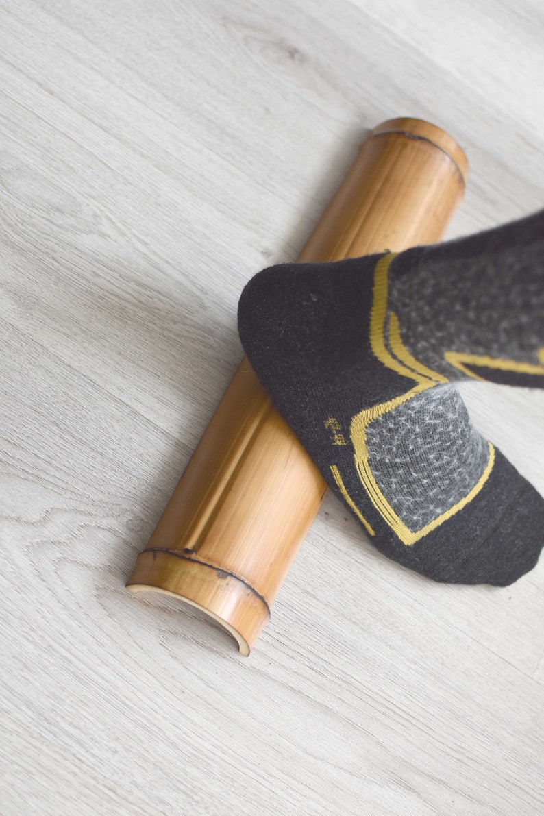 May include: A person's foot wearing a black and gray sock with yellow accents is rolling a round, natural bamboo roller on a light gray wood floor.