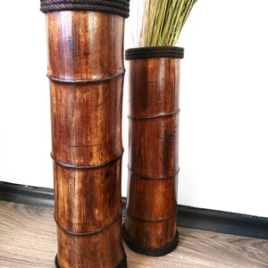 Puede incluir: Dos jarrones altos de bamb&uacute; marr&oacute;n con ribete de cuerda negra. Los jarrones est&aacute;n vac&iacute;os y est&aacute;n de pie sobre un suelo de madera.