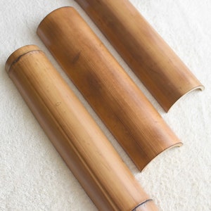 May include: Three natural brown bamboo rollers of varying sizes. The rollers are curved on one end and flat on the other. The rollers are arranged in a row on a white fluffy surface.
