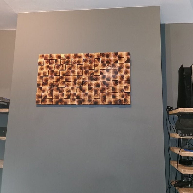 Op de afbeelding: Een wandkunstwerk gemaakt van houten blokken, gerangschikt in een geometrisch patroon. De blokken zijn donkerbruin van kleur en hebben een natuurlijke houtnerf. Het kunstwerk is gemonteerd op een grijze muur.