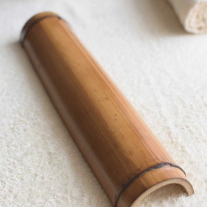 May include: A long, brown, cylindrical bamboo roller with a slightly curved end. The roller is lying on a white, fluffy surface.