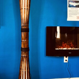 Op de afbeelding: Een hoge, bruine bamboe vloerlamp met een witte stoffen kap. De lamp is aan en geeft een warm licht. De lamp staat op een houten vloer met een blauwe muur op de achtergrond.