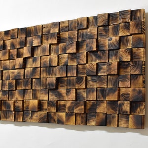 Op de afbeelding: Een wandkunstwerk gemaakt van gebrand houten vierkanten, gerangschikt in een willekeurig patroon. Het hout heeft een donkerbruine kleur met een licht getextureerd oppervlak.