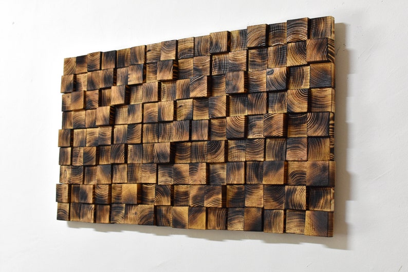 Op de afbeelding: Een wandkunstwerk gemaakt van gebrand houten vierkanten, gerangschikt in een willekeurig patroon. Het hout heeft een donkerbruine kleur met een licht getextureerd oppervlak.