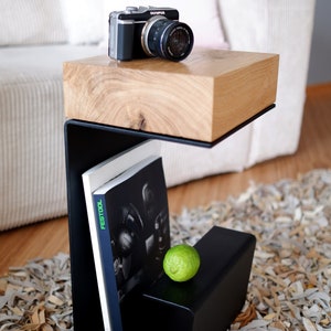 Op de afbeelding: Een zwarte metalen bijzettafel met een houten blad. De tafel heeft een plank voor boeken en een kleine ruimte voor een limoen. Een camera ligt op het tafelblad.