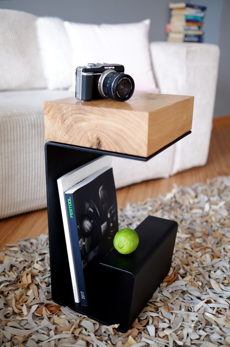 Op de afbeelding: Een zwarte metalen bijzettafel met een houten blad. De tafel heeft een plank voor boeken en een kleine ruimte voor een limoen. Een camera ligt op het tafelblad.