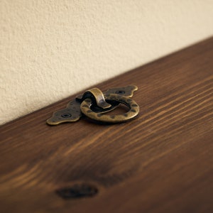 May include: A close-up of a brass ring pull handle on a wooden surface. The handle is attached to a decorative plate with a scalloped edge.