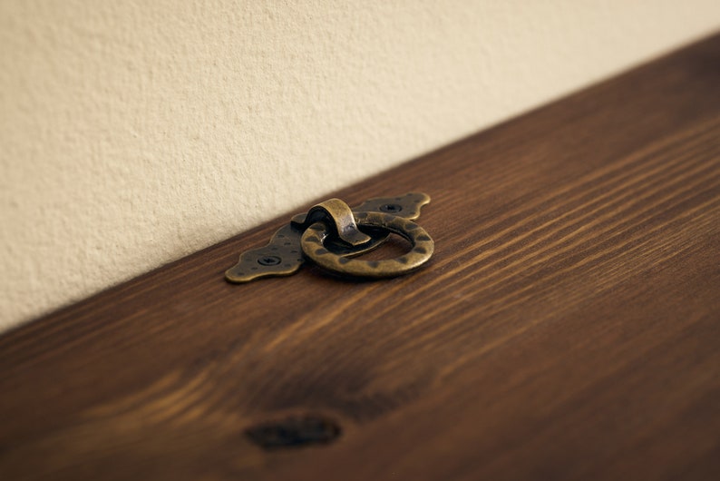 May include: A close-up of a brass ring pull handle on a wooden surface. The handle is attached to a decorative plate with a scalloped edge.