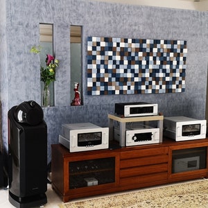 May include: A home theater system with a large wooden cabinet, multiple silver audio components, and two large black speakers. The system is set up in a room with a gray textured wall and a decorative wall panel made of small wooden squares.