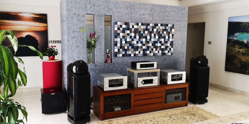 May include: A home theater system with a large wooden cabinet, multiple silver audio components, and two large black speakers. The system is set up in a room with a gray textured wall and a decorative wall panel made of small wooden squares.