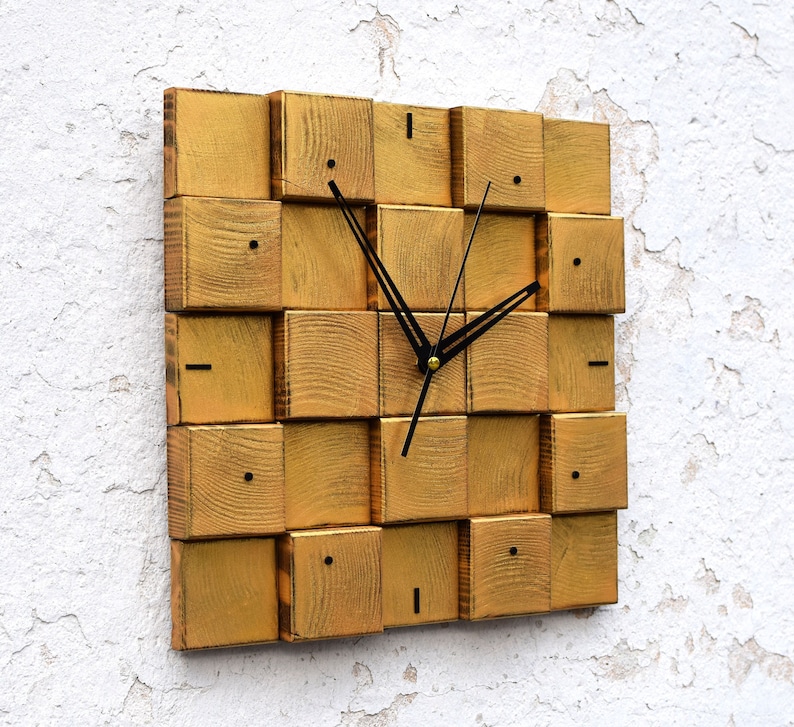 Puede incluir: Reloj de pared de madera en tono dorado con manecillas negras y dise&ntilde;o cuadrado. La esfera del reloj est&aacute; formada por cuadrados de madera individuales.