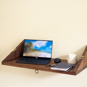 May include: A wooden wall-mounted desk with a laptop, a notebook, a pen, a mouse, and a white mug on top. The desk is folded up and has brass hinges.