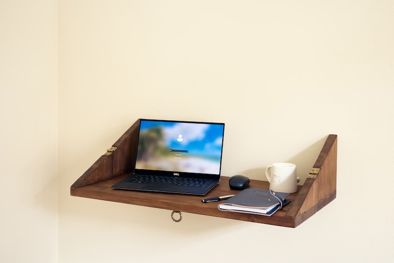 May include: A wooden wall-mounted desk with a laptop, a notebook, a pen, a mouse, and a white mug on top. The desk is folded up and has brass hinges.