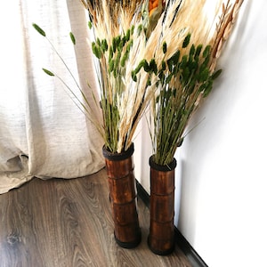 Puede incluir: Dos jarrones cil&iacute;ndricos altos y marrones hechos de bamb&uacute; est&aacute;n llenos de hierbas y flores secas. Los jarrones est&aacute;n sobre un suelo de madera frente a una pared blanca.