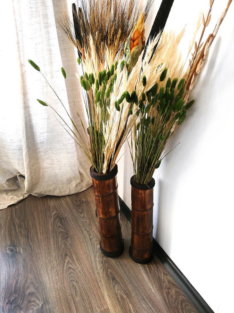 Puede incluir: Dos jarrones cil&iacute;ndricos altos y marrones hechos de bamb&uacute; est&aacute;n llenos de hierbas y flores secas. Los jarrones est&aacute;n sobre un suelo de madera frente a una pared blanca.