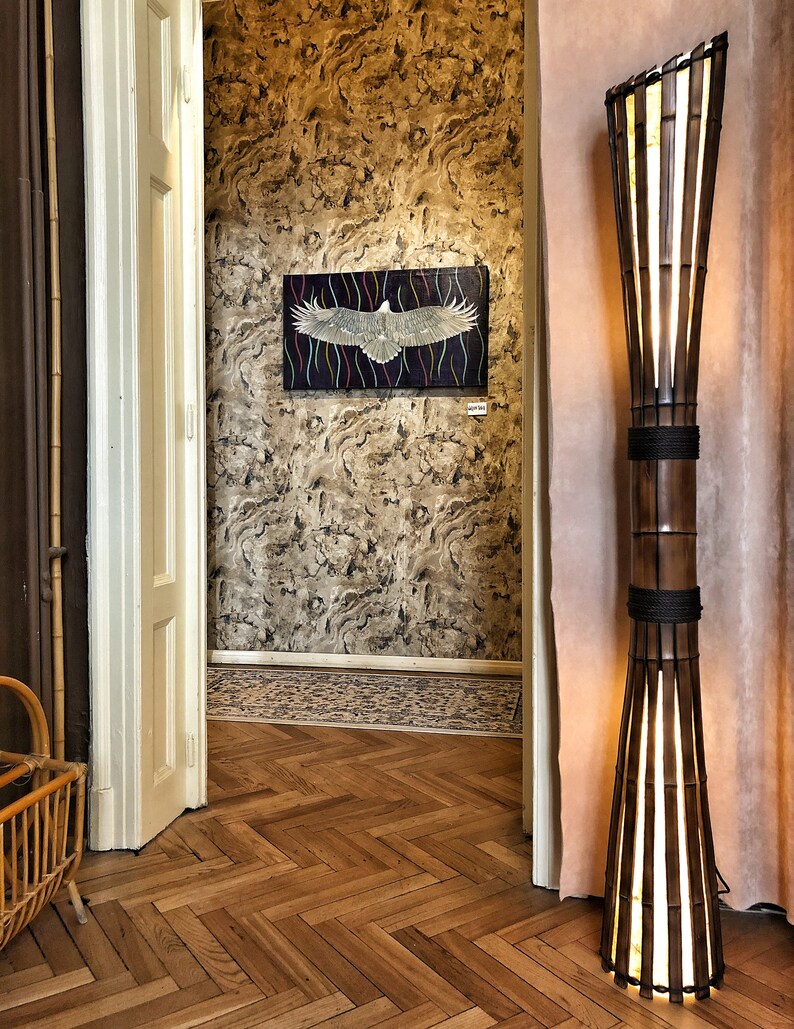 Op de afbeelding: Een hoge, houten vloerlamp met een lichtbruine afwerking en een geweven bamboe kap. De lamp staat op een houten vloer met een visgraatpatroon. De lamp bevindt zich in een kamer met een witte deur en een schilderij van een vogel aan de muur.