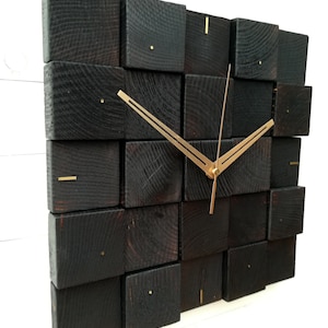 Puede incluir: Un reloj de pared de madera negra con manecillas doradas. La esfera del reloj est&aacute; formada por varios bloques de madera cuadrados.