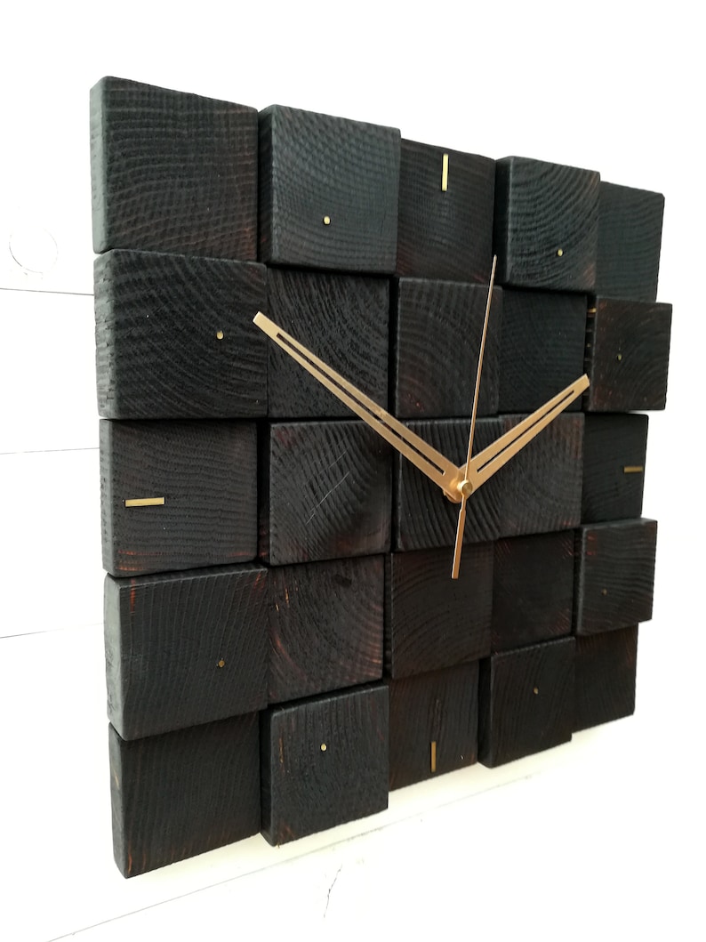 Puede incluir: Un reloj de pared de madera negra con manecillas doradas. La esfera del reloj est&aacute; formada por varios bloques de madera cuadrados.