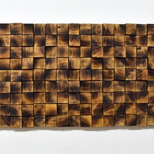 Op de afbeelding: Een wandkunstwerk gemaakt van gebrande houten blokken, gerangschikt in een geometrisch patroon. De blokken zijn in verschillende tinten bruin en zwart, wat een rustieke en moderne uitstraling cre&euml;ert.
