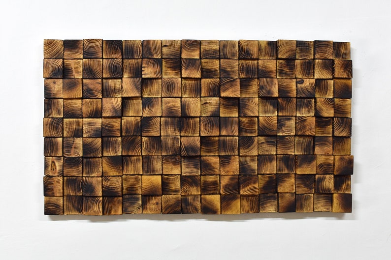 Op de afbeelding: Een wandkunstwerk gemaakt van gebrande houten blokken, gerangschikt in een geometrisch patroon. De blokken zijn in verschillende tinten bruin en zwart, wat een rustieke en moderne uitstraling cre&euml;ert.