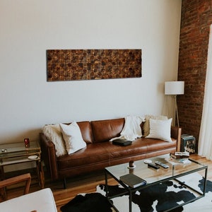 Puede incluir: Un sof&aacute; de cuero marr&oacute;n con almohadas blancas se encuentra en una sala de estar con una pieza de arte de pared de madera marr&oacute;n por encima. La habitaci&oacute;n tiene una mesa de caf&eacute; de vidrio, una alfombra de piel de vaca blanca y una l&aacute;mpara de pie.