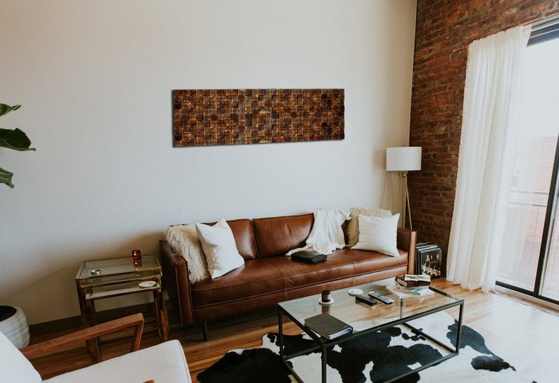 Puede incluir: Un sof&aacute; de cuero marr&oacute;n con almohadas blancas se encuentra en una sala de estar con una pieza de arte de pared de madera marr&oacute;n por encima. La habitaci&oacute;n tiene una mesa de caf&eacute; de vidrio, una alfombra de piel de vaca blanca y una l&aacute;mpara de pie.