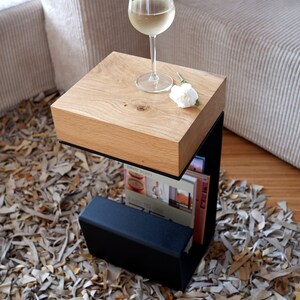 Op de afbeelding: Een houten bijzettafel met een zwart metalen frame en een plank om boeken of tijdschriften op te bergen. Op het tafelblad staat een glas witte wijn en een witte bloem.