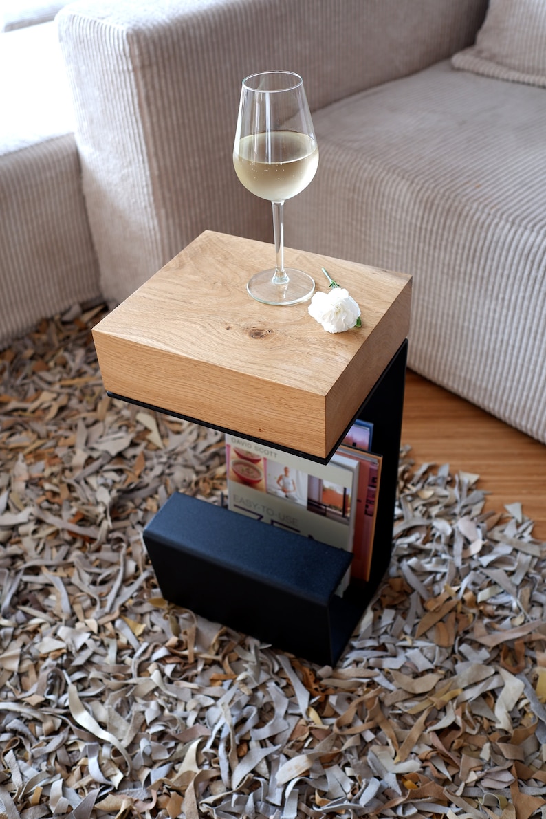 Op de afbeelding: Een houten bijzettafel met een zwart metalen frame en een plank om boeken of tijdschriften op te bergen. Op het tafelblad staat een glas witte wijn en een witte bloem.