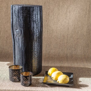 May include: A black charred wood stump with a natural wood grain pattern, used as a side table. Two gold metal candle holders with a cut-out design and three yellow lemons on a black square plate are on a brown surface.