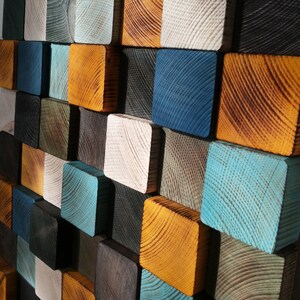 Puede incluir: Una pieza de arte de pared hecha de cuadrados de madera en varios colores, incluyendo azul, verde, marr&oacute;n y amarillo. Los cuadrados est&aacute;n dispuestos en un patr&oacute;n escalonado, creando una superficie visualmente interesante y texturizada.