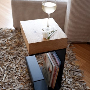 Op de afbeelding: Een houten bijzettafel met een zwart metalen frame en een plank voor boeken. Een glas witte wijn en een witte bloem staan op het tafelblad.