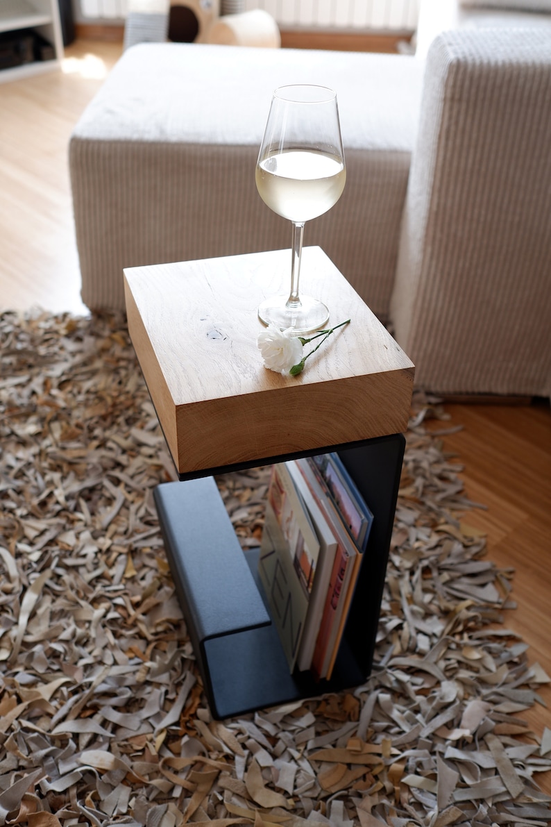 Op de afbeelding: Een houten bijzettafel met een zwart metalen frame en een plank voor boeken. Een glas witte wijn en een witte bloem staan op het tafelblad.