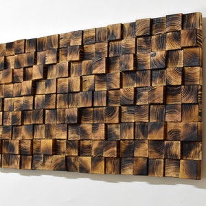 Puede incluir: Una pieza de arte de pared hecha de cuadrados de madera quemada, dispuestos en un patr&oacute;n aleatorio. La madera tiene un color marr&oacute;n oscuro con una superficie ligeramente texturizada.