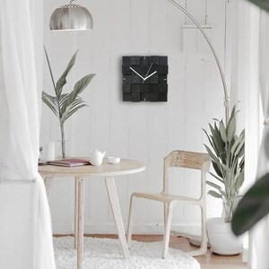 Puede incluir: Un reloj cuadrado negro con manecillas blancas cuelga de una pared blanca en una sala de estar moderna. El reloj est&aacute; hecho de bloques de madera y tiene un dise&ntilde;o minimalista. Una mesa blanca con una silla blanca se encuentra en primer plano.