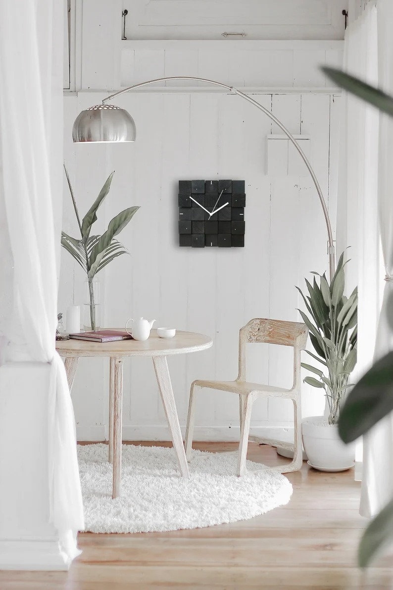 Puede incluir: Un reloj cuadrado negro con manecillas blancas cuelga de una pared blanca en una sala de estar moderna. El reloj est&aacute; hecho de bloques de madera y tiene un dise&ntilde;o minimalista. Una mesa blanca con una silla blanca se encuentra en primer plano.