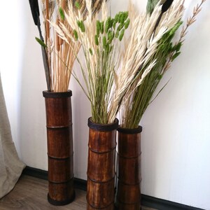 May include: Three tall brown bamboo vases with a dark brown stain, each holding dried grasses and flowers. The vases are arranged in a row on a wooden floor.