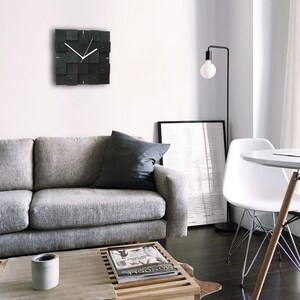 Puede incluir: Un sof&aacute; gris con un coj&iacute;n negro se encuentra en una sala de estar con un reloj blanco y negro en la pared, una mesa de caf&eacute; de madera con una revista encima, una silla blanca y una l&aacute;mpara de pie con una bombilla blanca.