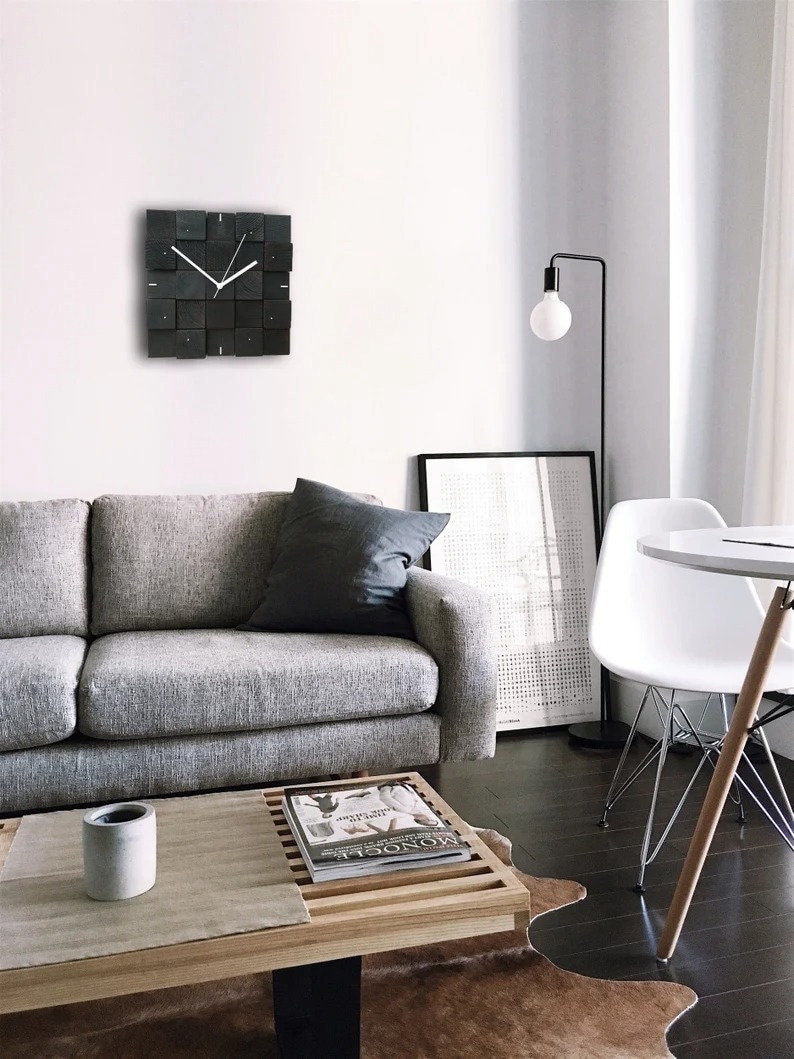Puede incluir: Un sof&aacute; gris con un coj&iacute;n negro se encuentra en una sala de estar con un reloj blanco y negro en la pared, una mesa de caf&eacute; de madera con una revista encima, una silla blanca y una l&aacute;mpara de pie con una bombilla blanca.