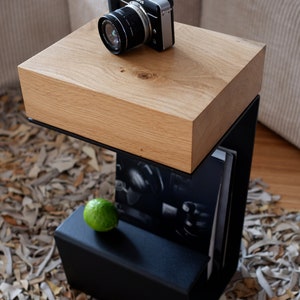Op de afbeelding: Een zwarte metalen bijzettafel met een houten blad. De tafel heeft een plank voor opslag en staat op een tapijt gemaakt van leren stroken.